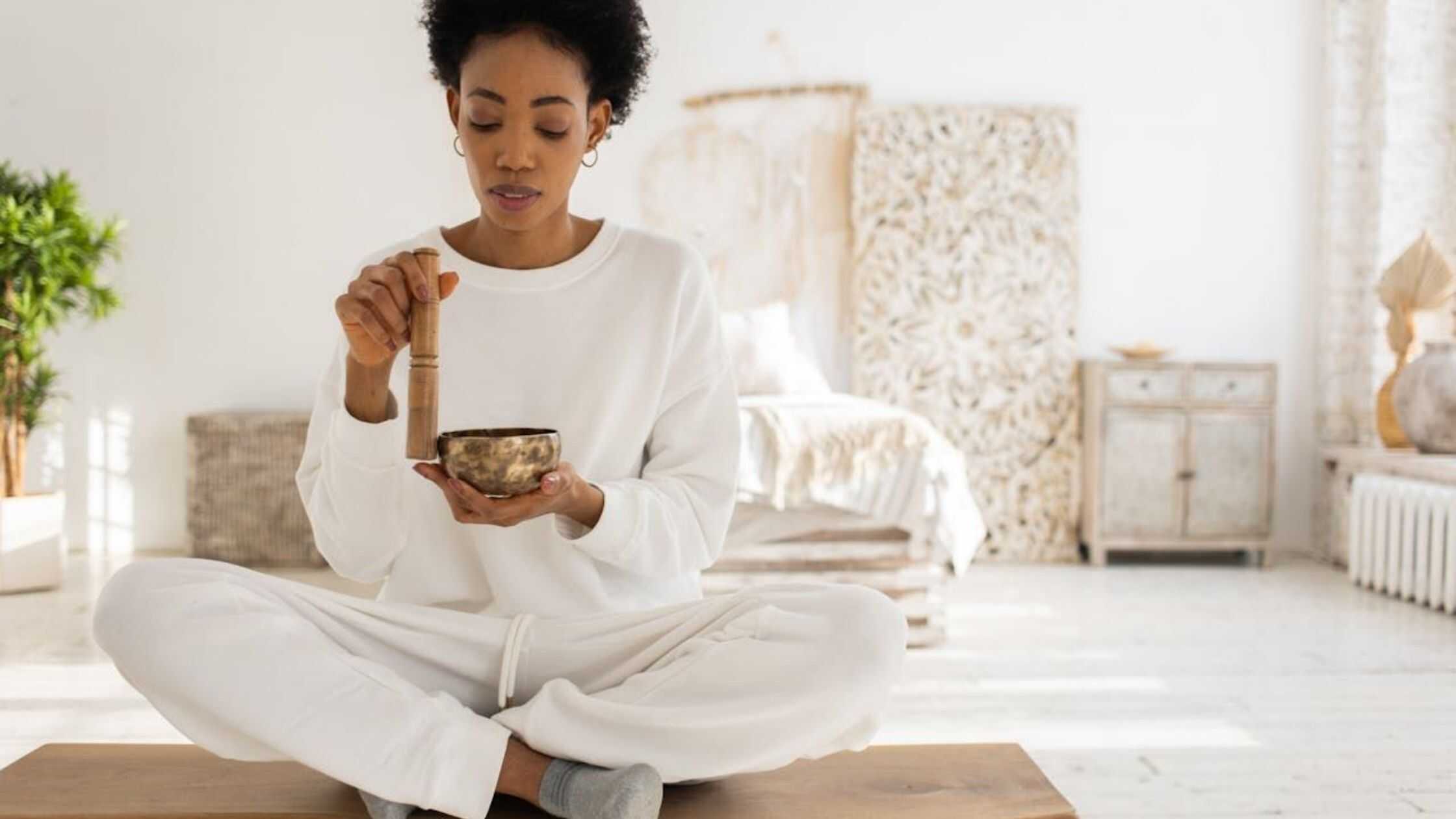 Peaceful woman meditating and playing singing bowl for relaxation and spiritual healing in a bright cozy bedroom.